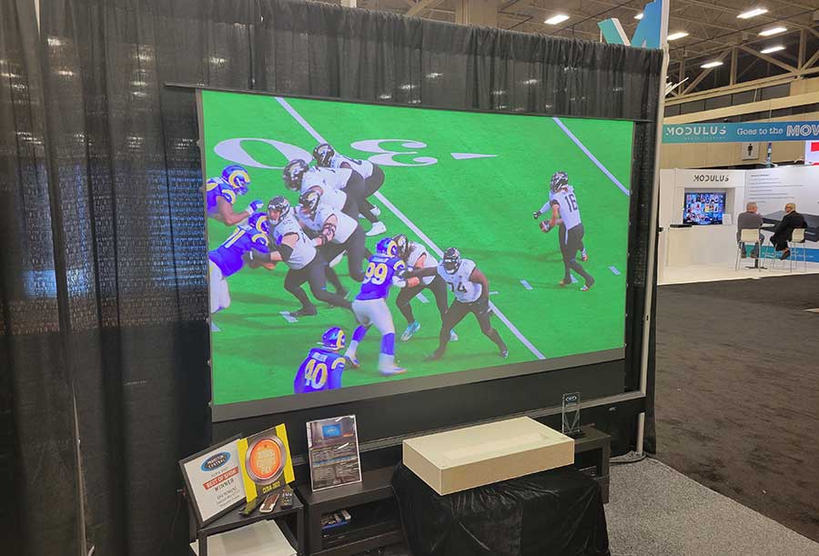 Large screen displaying a football game in an indoor setting with booths and people in the background.