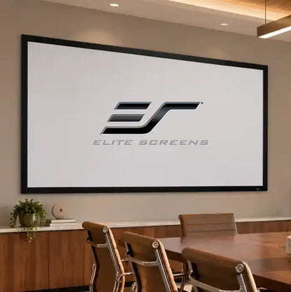 A large wall-mounted projection screen in a modern conference room setting, displaying the Elite Screens logo. The screen is framed in black and hangs on a beige wall above a wooden table and chairs.