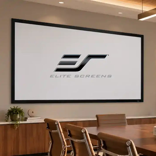 A large wall-mounted projection screen in a modern conference room setting, displaying the Elite Screens logo. The screen is framed in black and hangs on a beige wall above a wooden table and chairs.