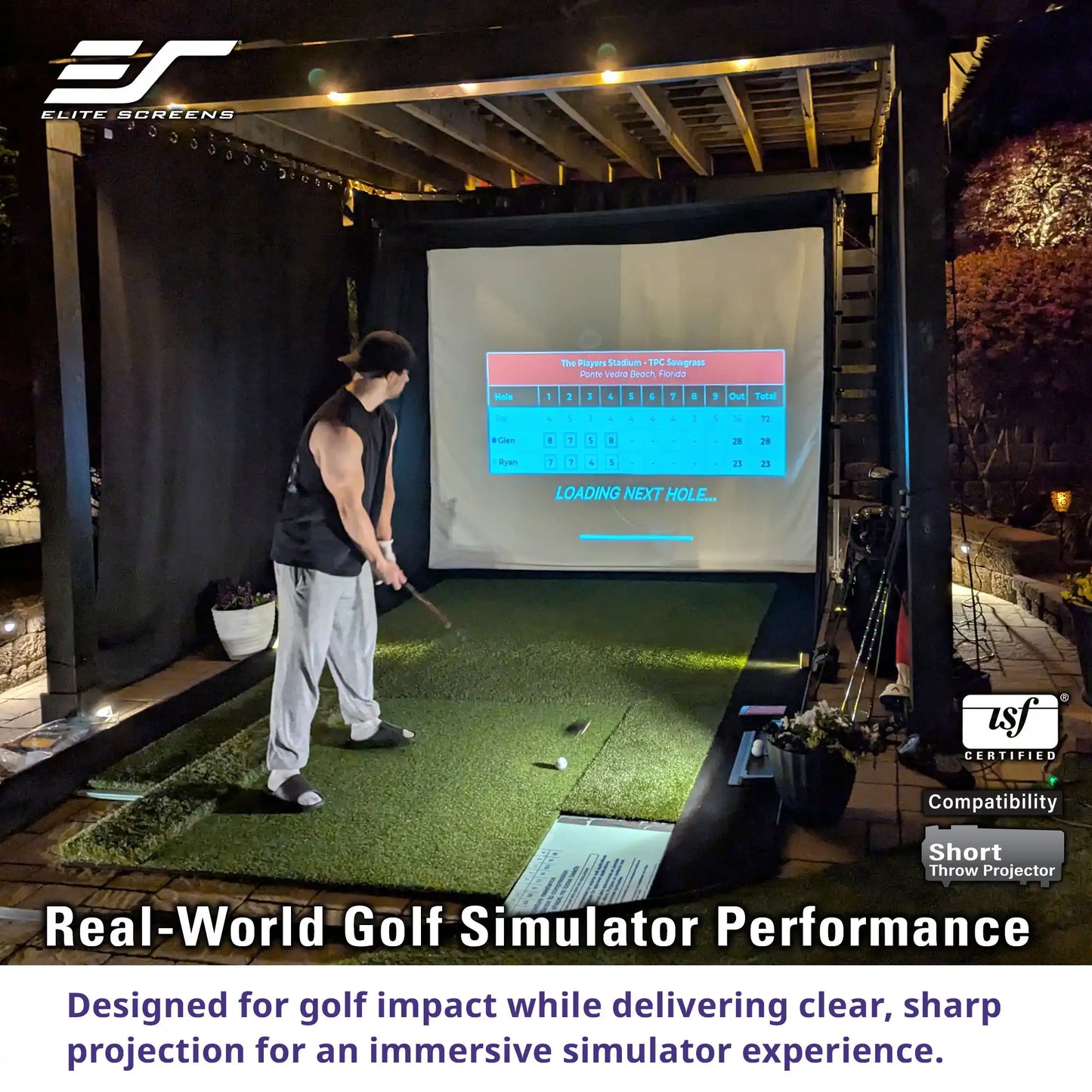 A man in a black sleeveless shirt and light gray sweatpants is playing a golf simulator at night. The simulator setup includes a green turf hitting surface, a large projection screen displaying a golf course layout, and a black enclosure.