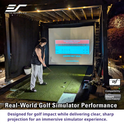 A man in a black sleeveless shirt and light gray sweatpants is playing a golf simulator at night. The simulator setup includes a green turf hitting surface, a large projection screen displaying a golf course layout, and a black enclosure.