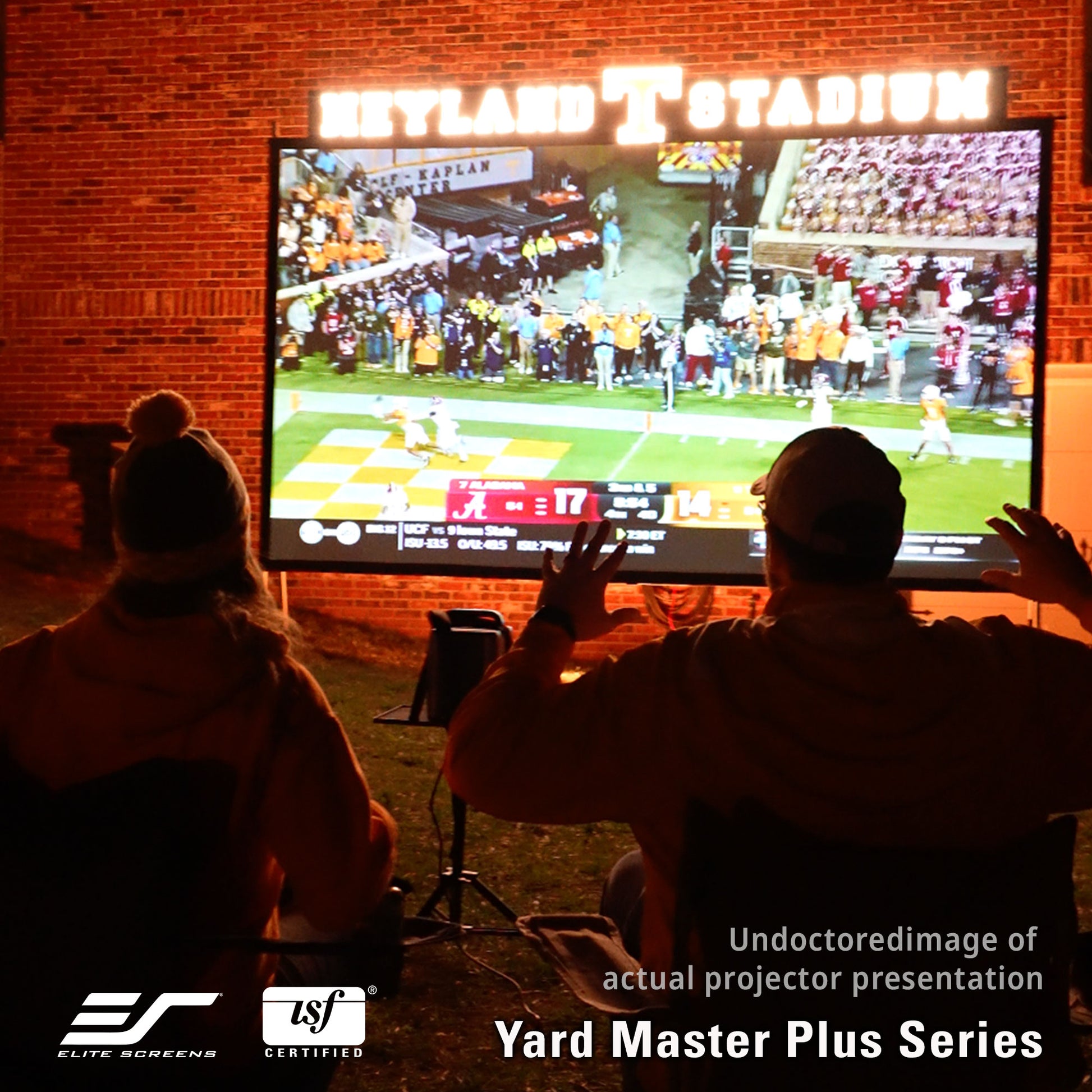 Two people watching a sports game projected onto a screen outdoors with 'Elite Screens' and 'TSF Certified' logos.